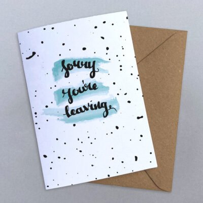 Leaving card with sorry you're leaving written in calligraphy on top of a blue watercolour swipe on a black splattered background
