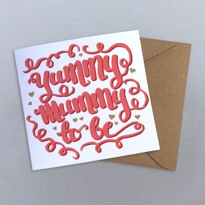 A hand calligraphy greeting card with yummy mummy to be in coral pink, surrounded by swirls and gold hearts on a white background