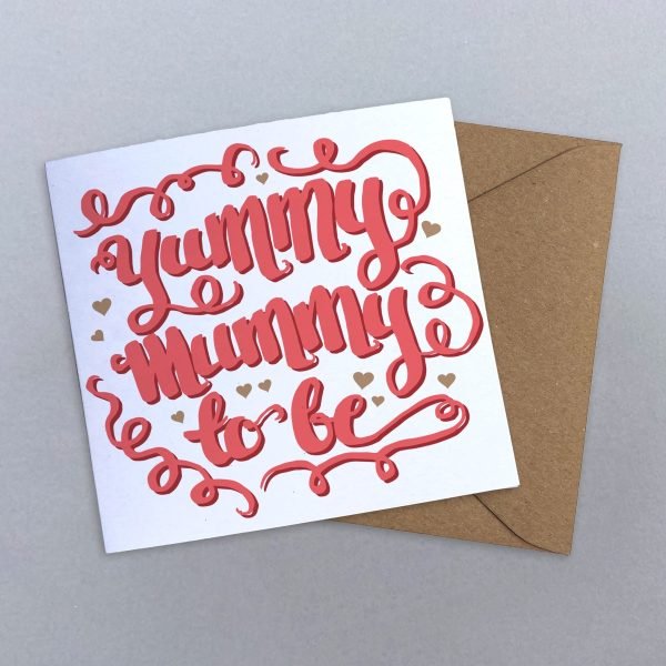 A hand calligraphy greeting card with yummy mummy to be in coral pink, surrounded by swirls and gold hearts on a white background